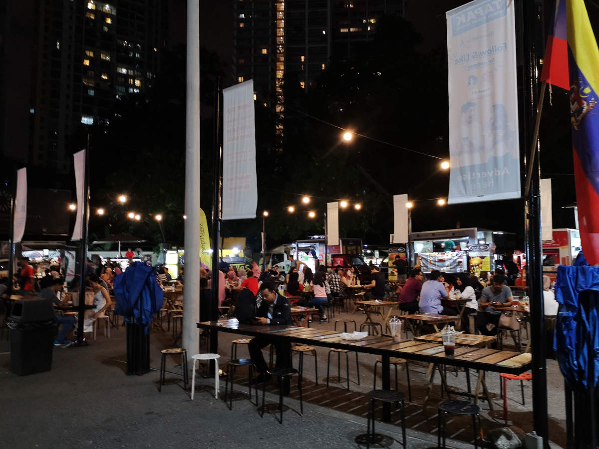 Außen herum stehen die Foodtrucks im Kreis, innen eine große Anzahl an Tischen und Stühlen, wo man bequem essen und trinken kann. Tapak Street Food Market in Kuala Lumpur KLCC. Wanderhunger