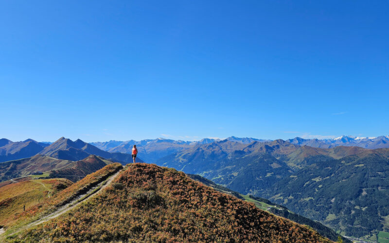 Frau in organge-farbenem Shirt auf Bergspitze umgeben von Bergpanorama